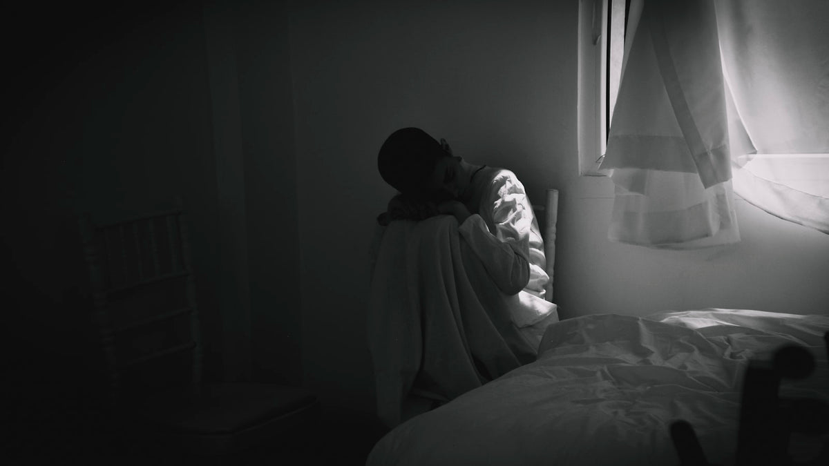 Person in a grey hoodie sitting on the edge of a bed in soft morning light