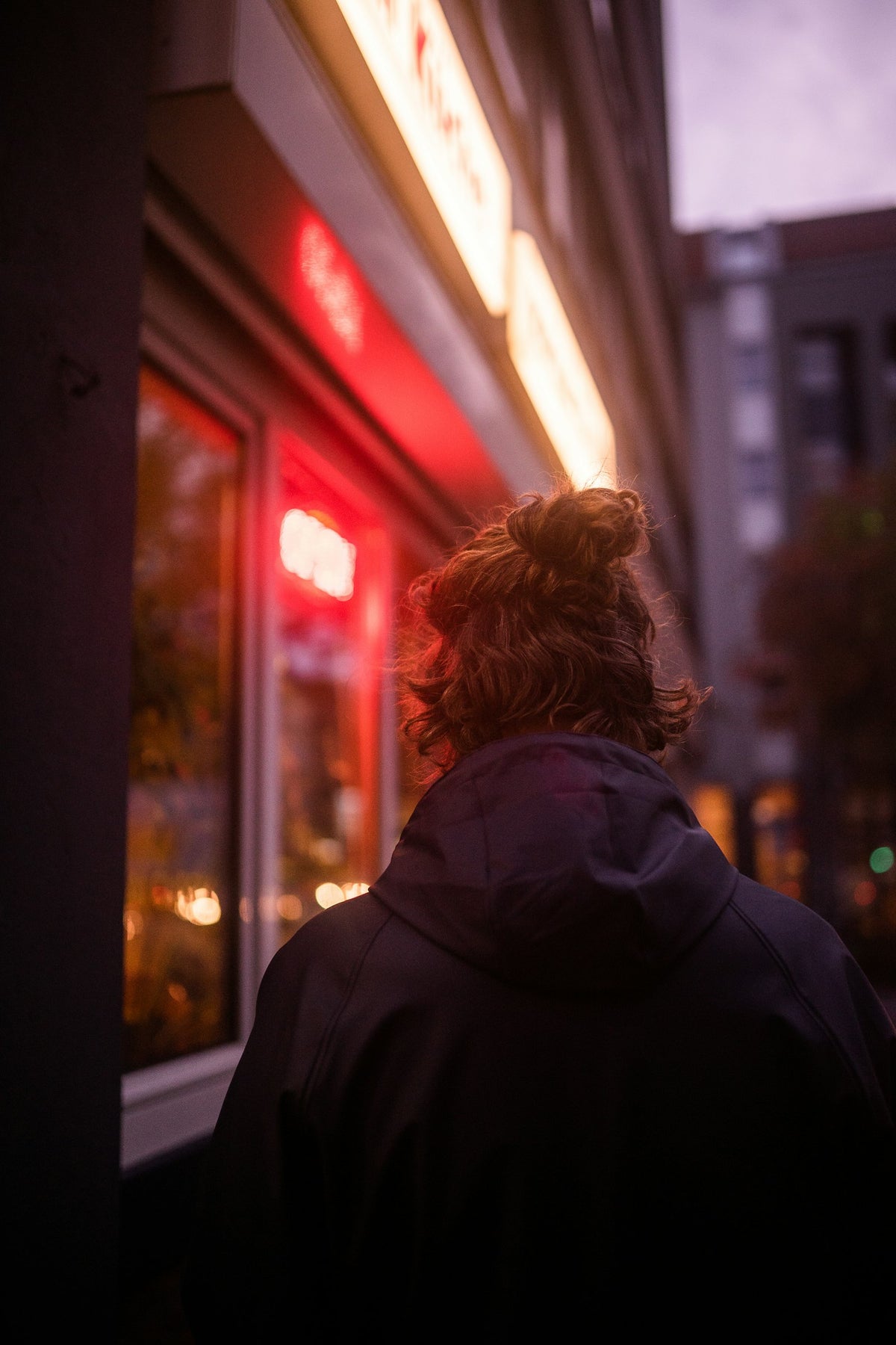 A student in an oversized black hoodie standing by a glass window at night, city lights blurred behind her