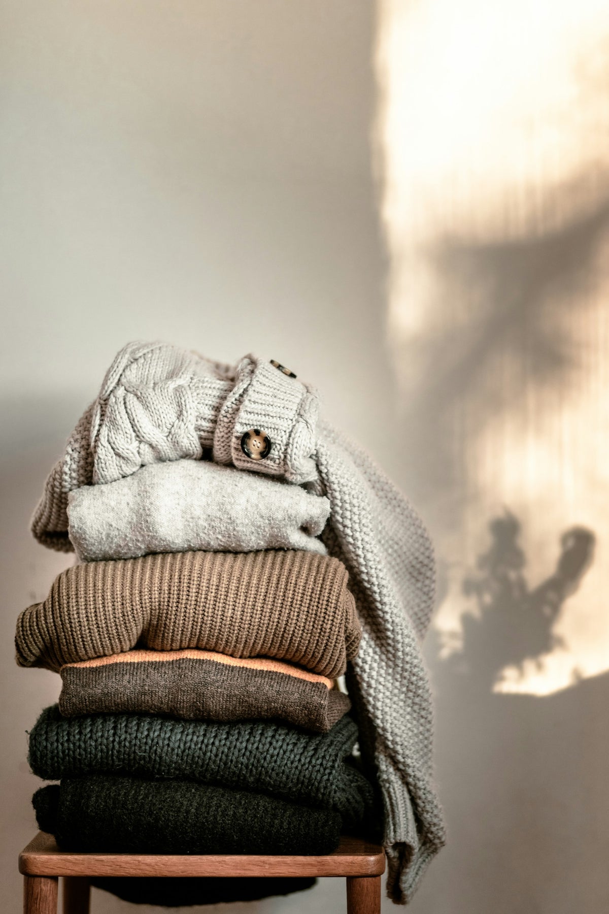 A stack of soft folded knitted hoodies and sweaters on a wooden chair in warm light