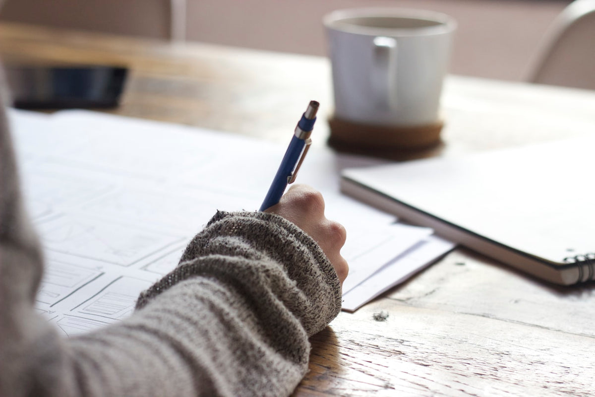 Person writing at a warm wooden desk with a ceramic mug