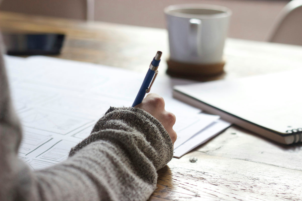 Person writing on a warm wooden desk with a coffee mug — student study scene