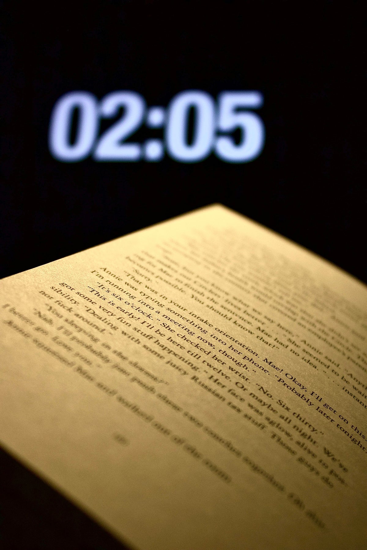Open book beside a digital clock showing 02:05 in a dimly lit room